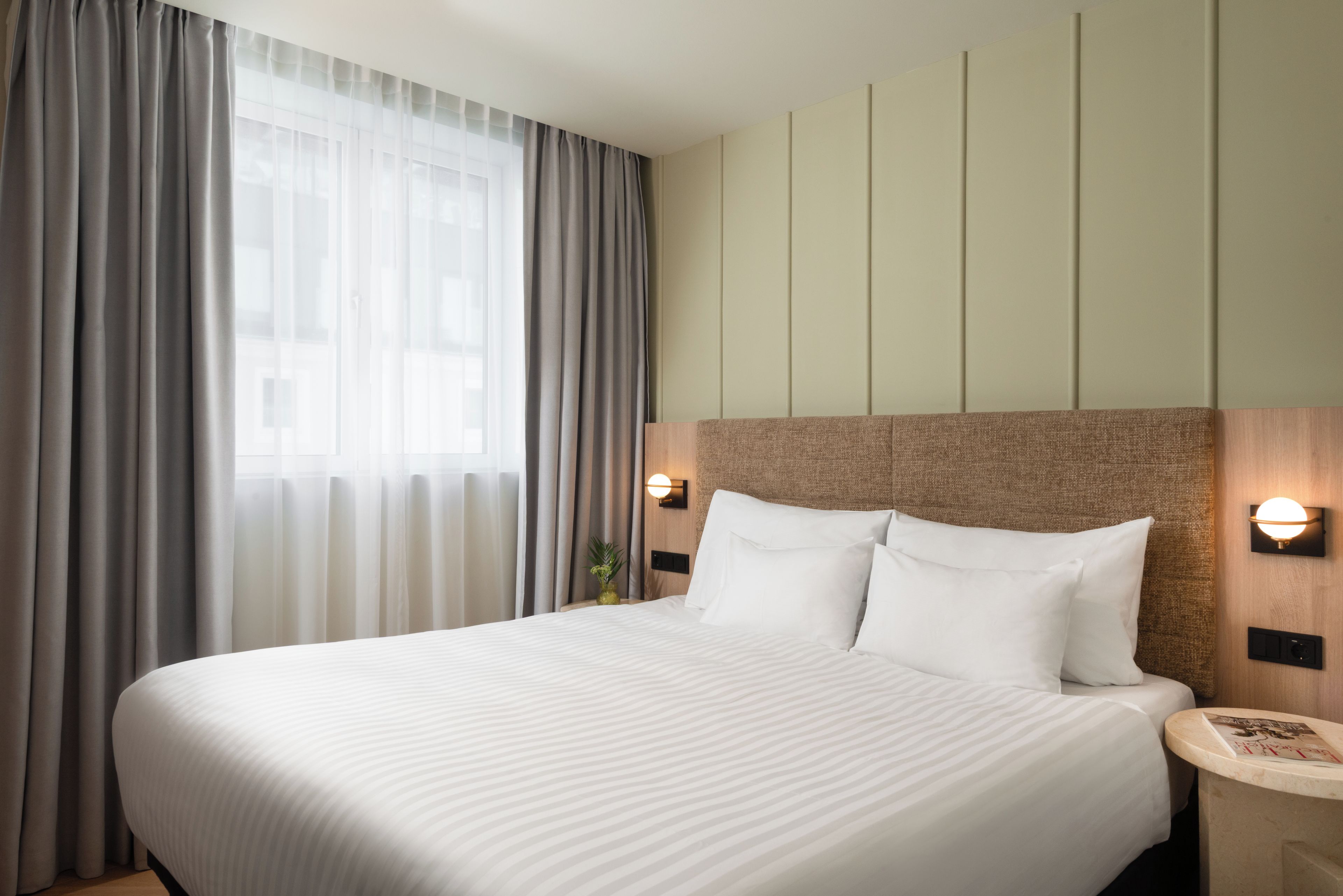 A hotel room with a large bed, a nightstand, a wall-mounted lamp, and a window with curtains.