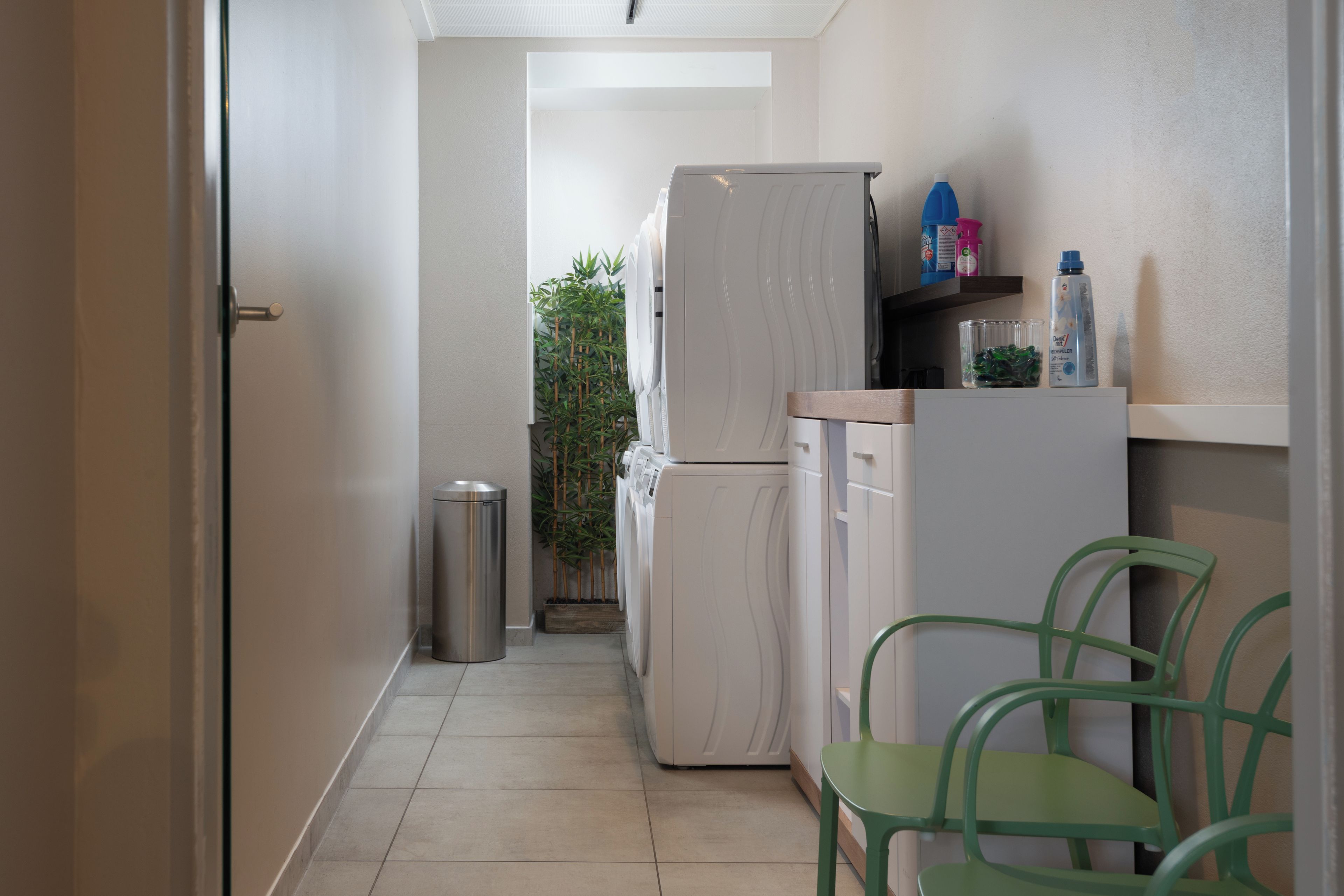A compact kitchen with a refrigerator, washing machine, and a trash bin. There are chairs and a shelf with cleaning supplies.