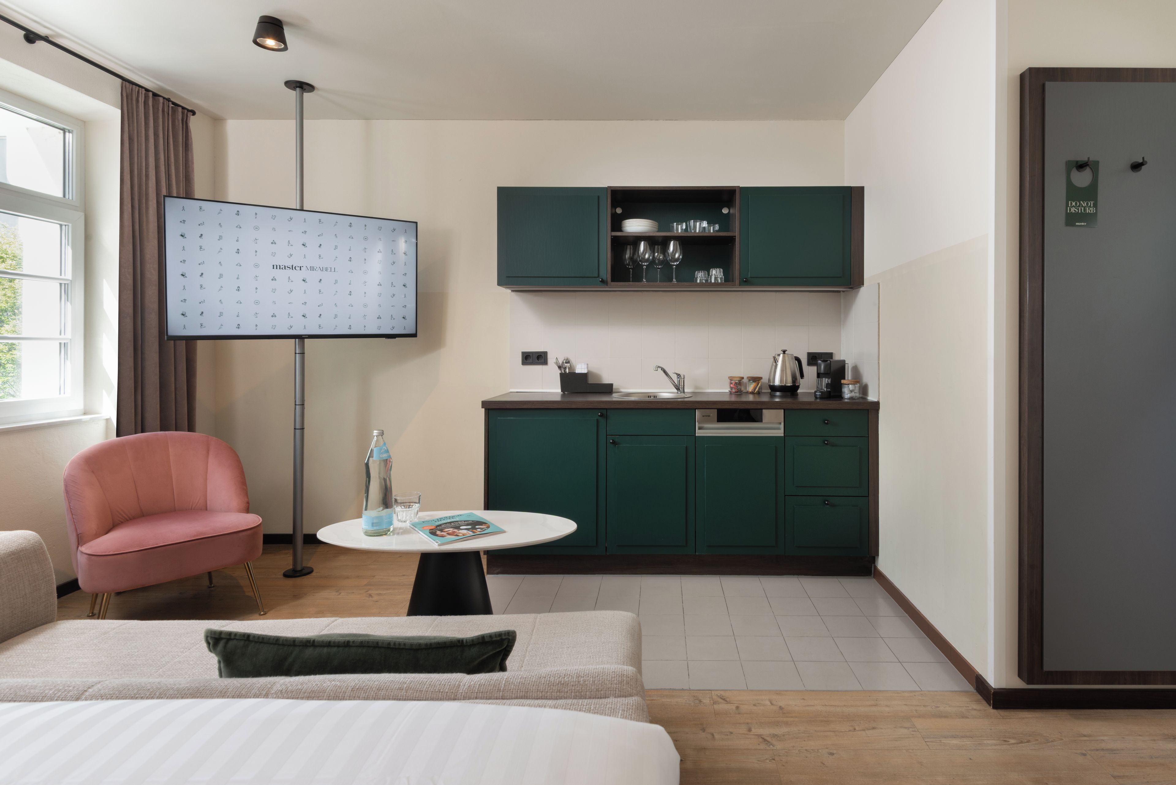 Habitació d'hotel moderna amb llit, taula rodona petita, cadira rosa, cuineta amb armaris verds i finestra gran.