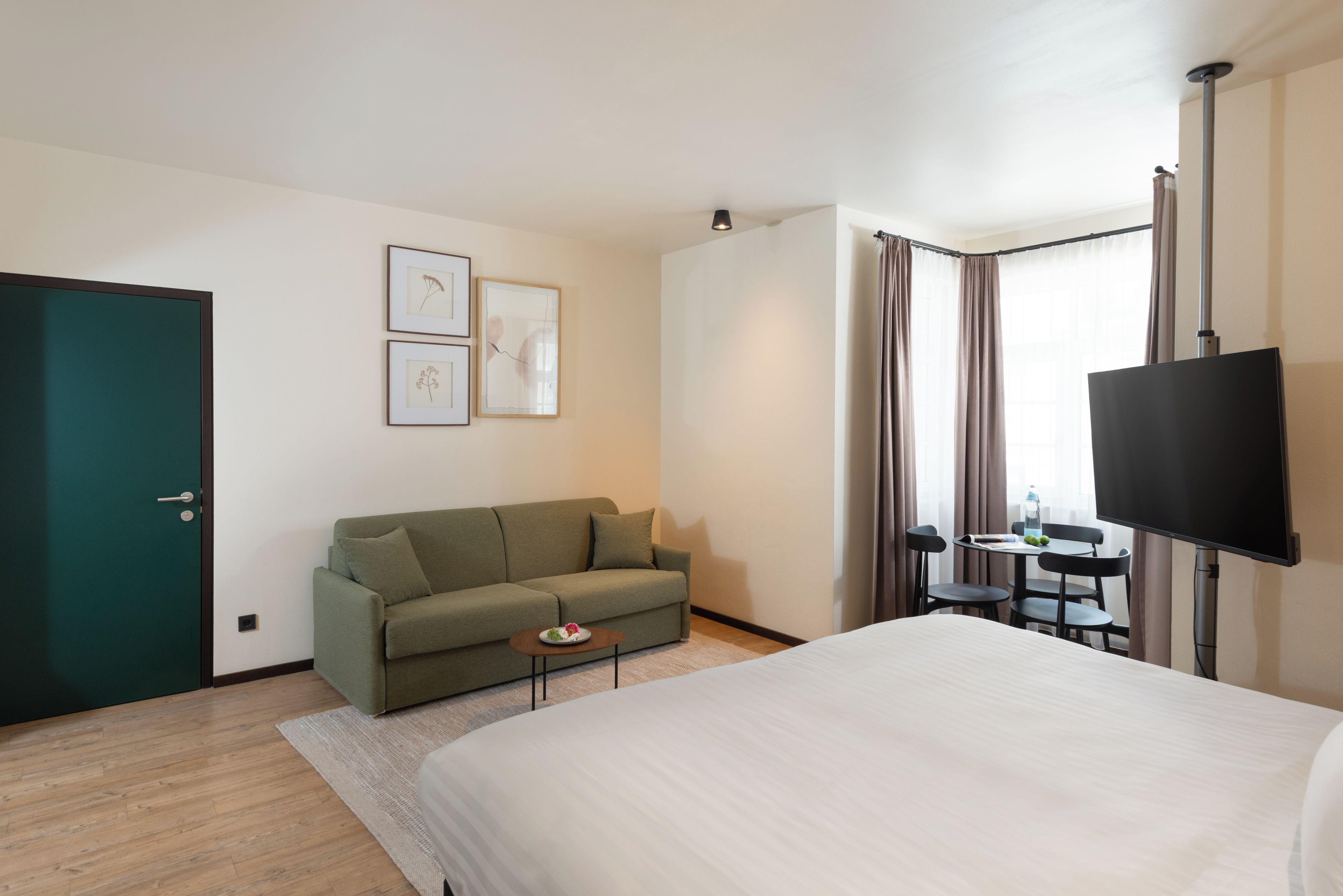 Ett hotellrum med en säng, en soffa, ett litet bord med en tallrik frukt, en tv och ett fönster med gardiner.
