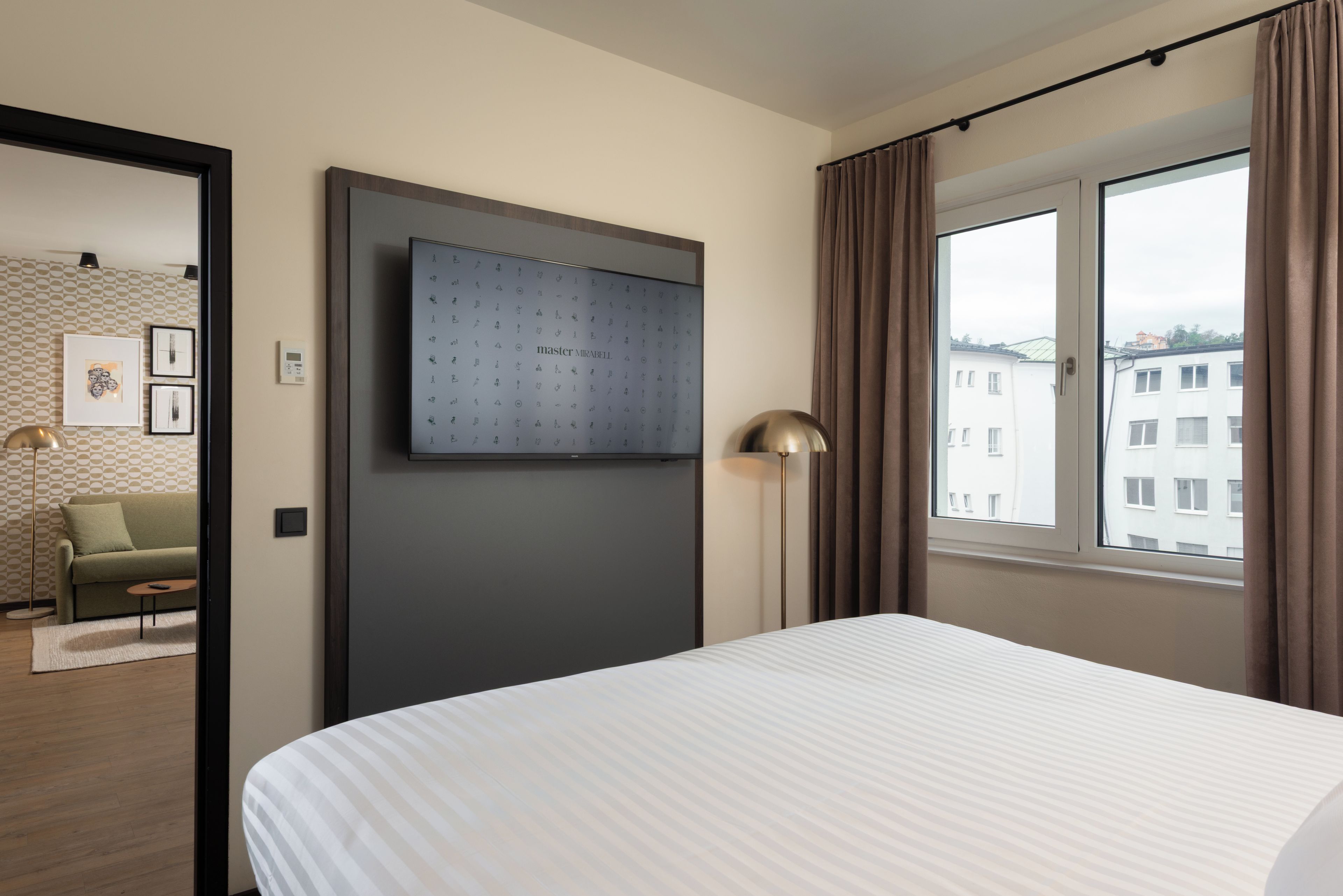 Habitación de hotel con cama, televisor montado en la pared, ventana con cortinas y vistas a edificios.