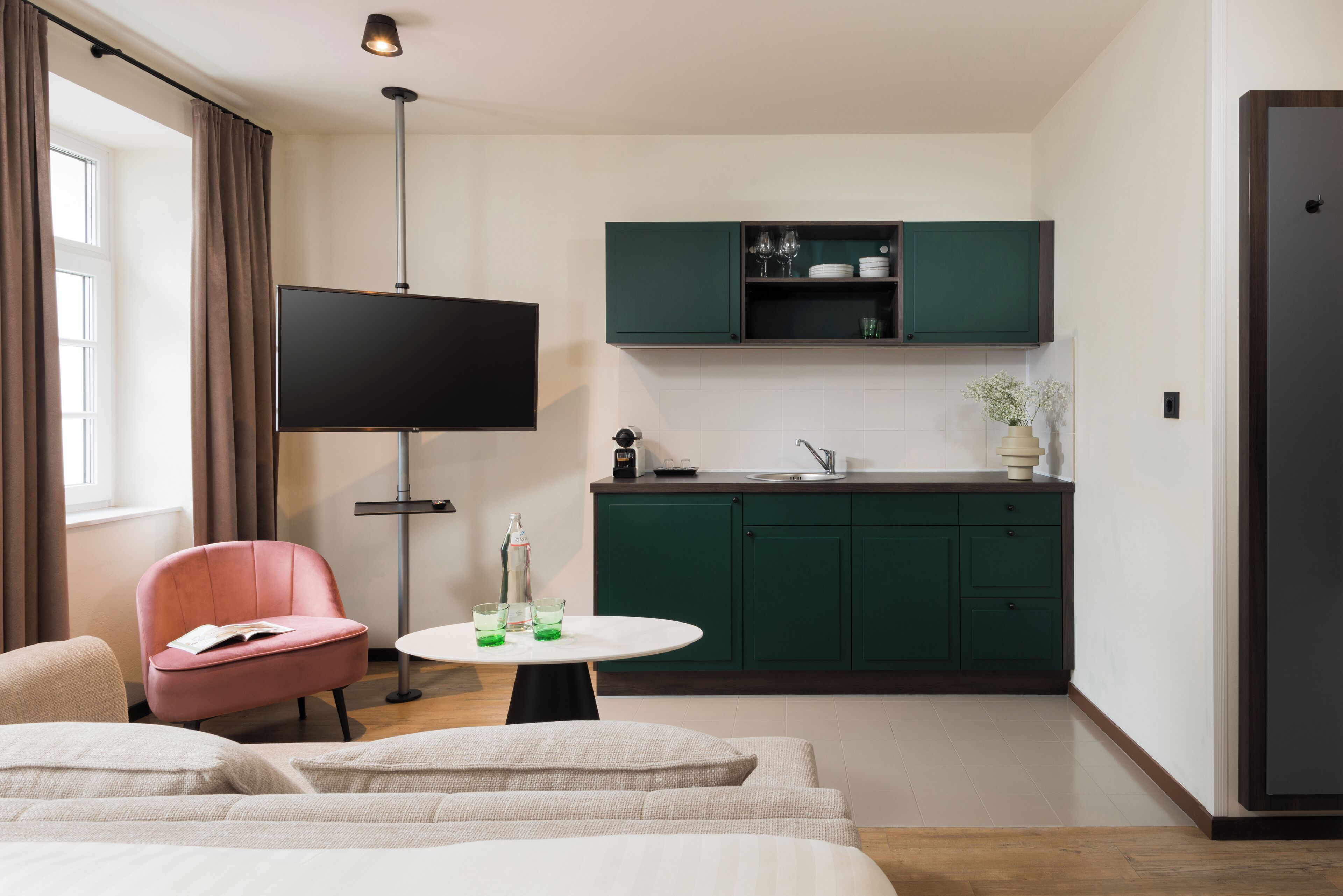 Habitación de hotel moderna con televisión de pantalla plana, una mesa de comedor con vasos verdes, una silla rosa y una cama con ropa de cama blanca.