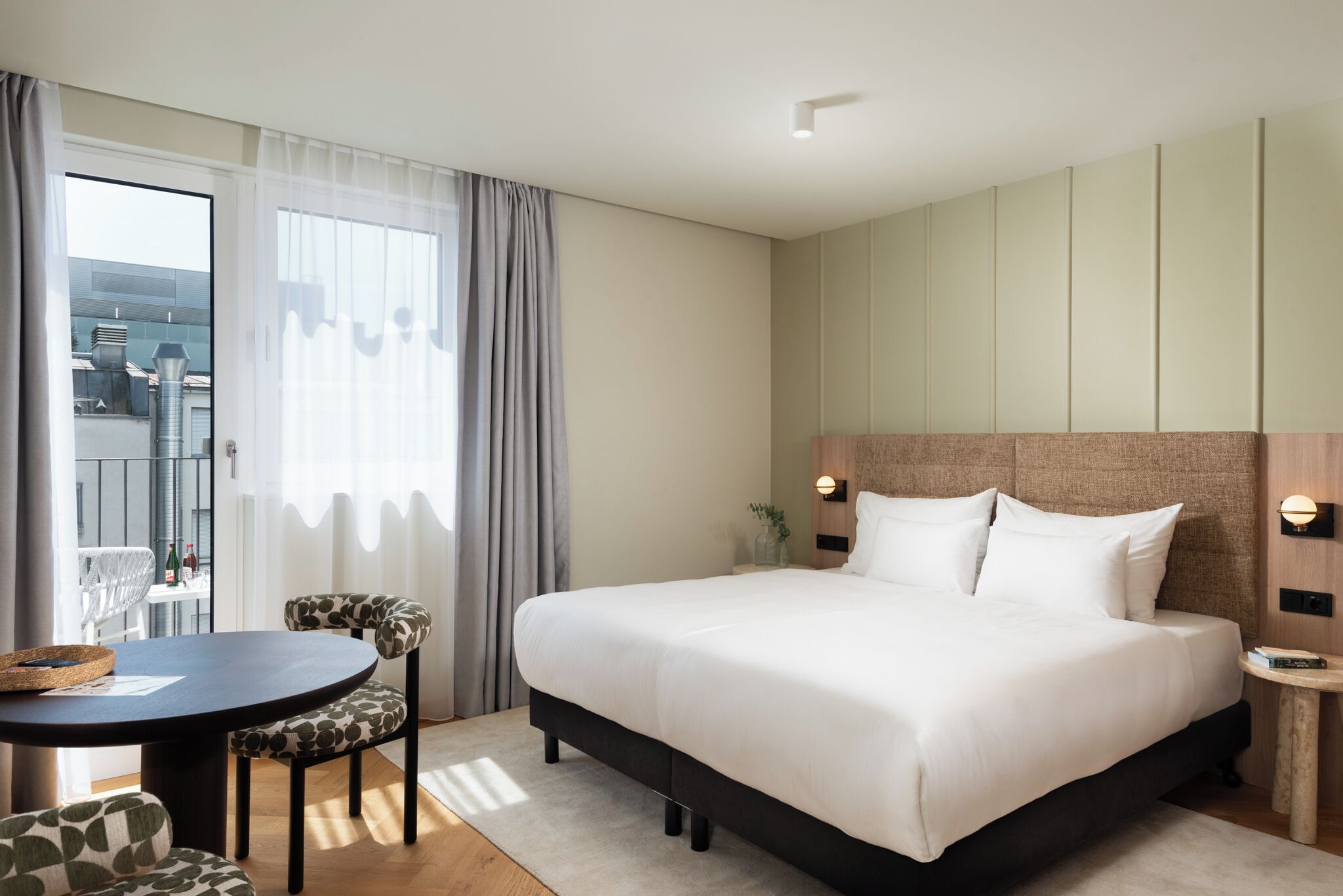 A hotel room with a large bed, a round dining table, and a view of the city through the window.