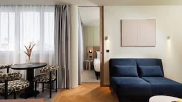 Una habitación de hotel con un sofá azul oscuro, una mesa redonda con sillas estampadas, una mesita auxiliar con un jarrón y una cama con cabecera.