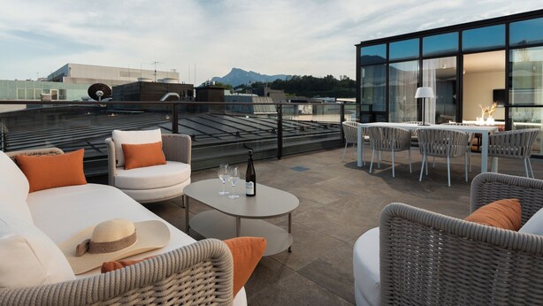 A rooftop terrace with wicker furniture, a table with a bottle and glasses, and a view of mountains. - master Mirabell (Salzburg)