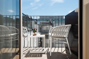Un balcón con dos sillones blancos de mimbre, una mesita con botellas y vasos, y vista a un edificio moderno.