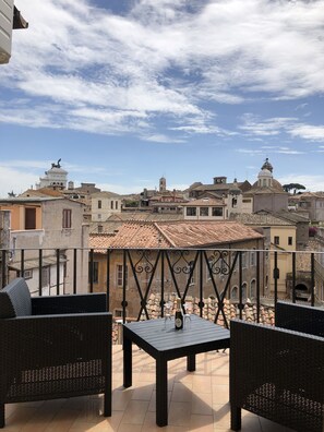 Une terrasse dotée de meubles en osier et offrant une vue sur une ville historique, avec ses bâtiments et son clocher.
