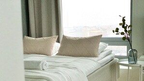 A neatly made bed with a white duvet and two pillows, a small bedside table with a vase of greenery, and a window with a view of buildings. - City Center Tower Apartment (Tampere)
