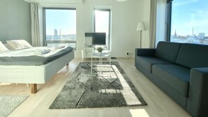 A modern hotel room with a bed, a sofa, a TV, and a large window offering a city view. - City Center Tower Apartment (Tampere)