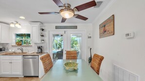 Une cuisine moderne avec des armoires blanches, une table à manger entourée de chaises en osier, un ventilateur de plafond et une vue sur l’extérieur grâce à une porte vitrée.