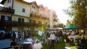 An outdoor event with people gathered around tables and chairs, a green tent, and a building with balconies. - Hotel Olimpic (Baselga di Pine)