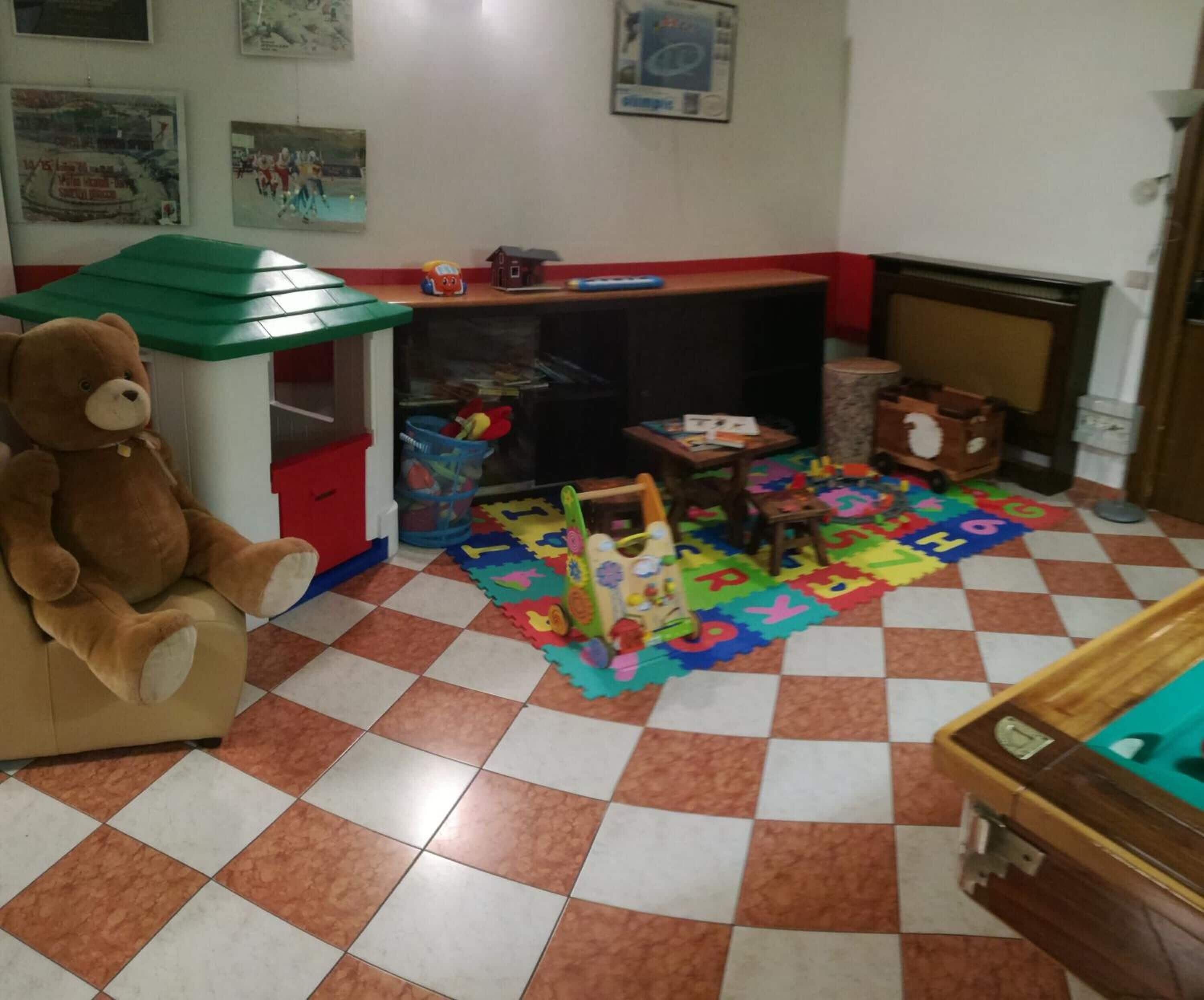 Una sala giochi con un grande orsacchiotto, un tavolino da gioco, puzzle dell'alfabeto e un trenino giocattolo.