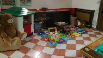Una sala giochi con un grande orsacchiotto, un tavolino da gioco, puzzle dell'alfabeto e un trenino giocattolo.
