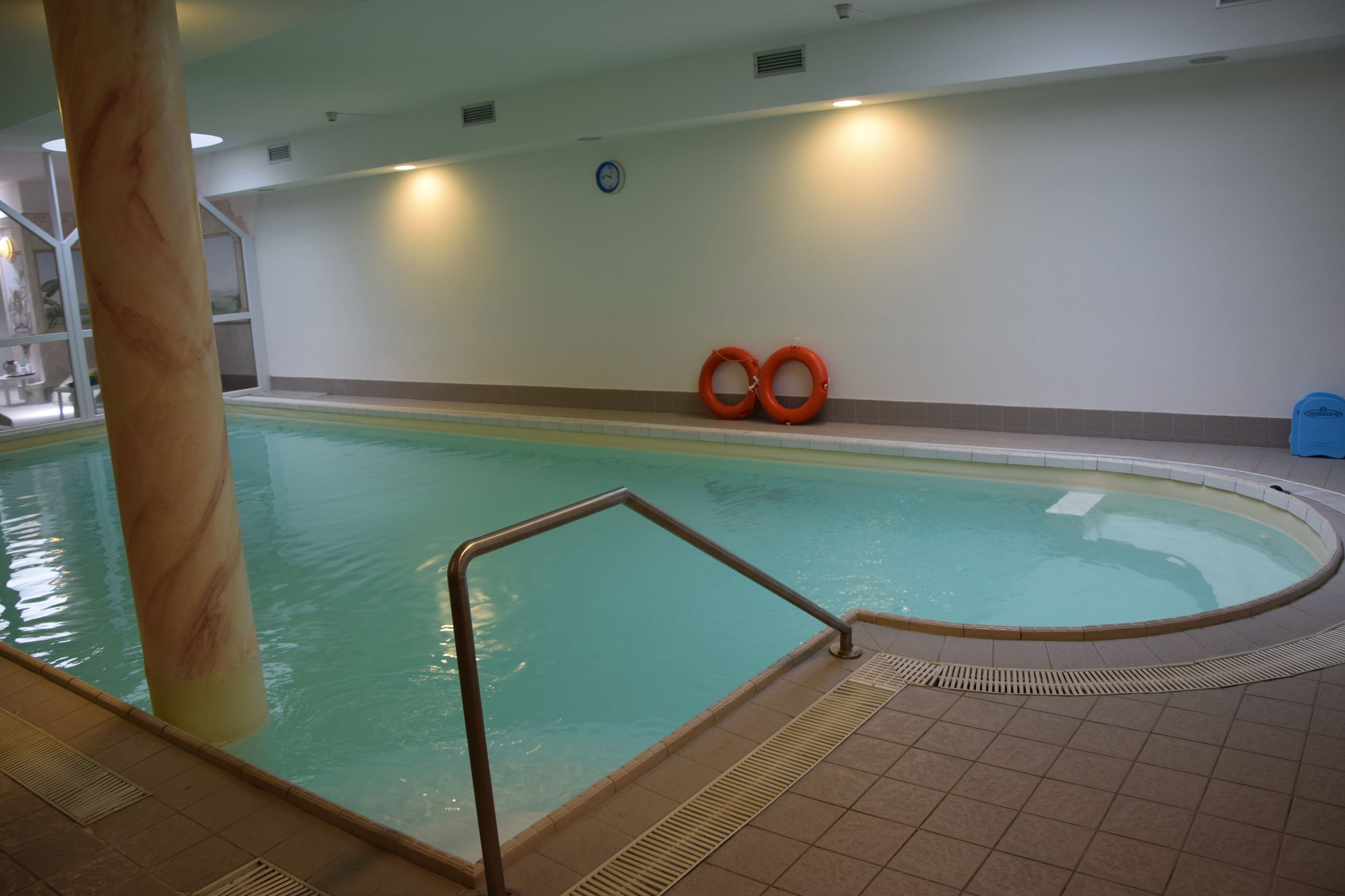 An indoor swimming pool with a tiled floor, a metal pool ladder, and two orange life rings on the wall.
