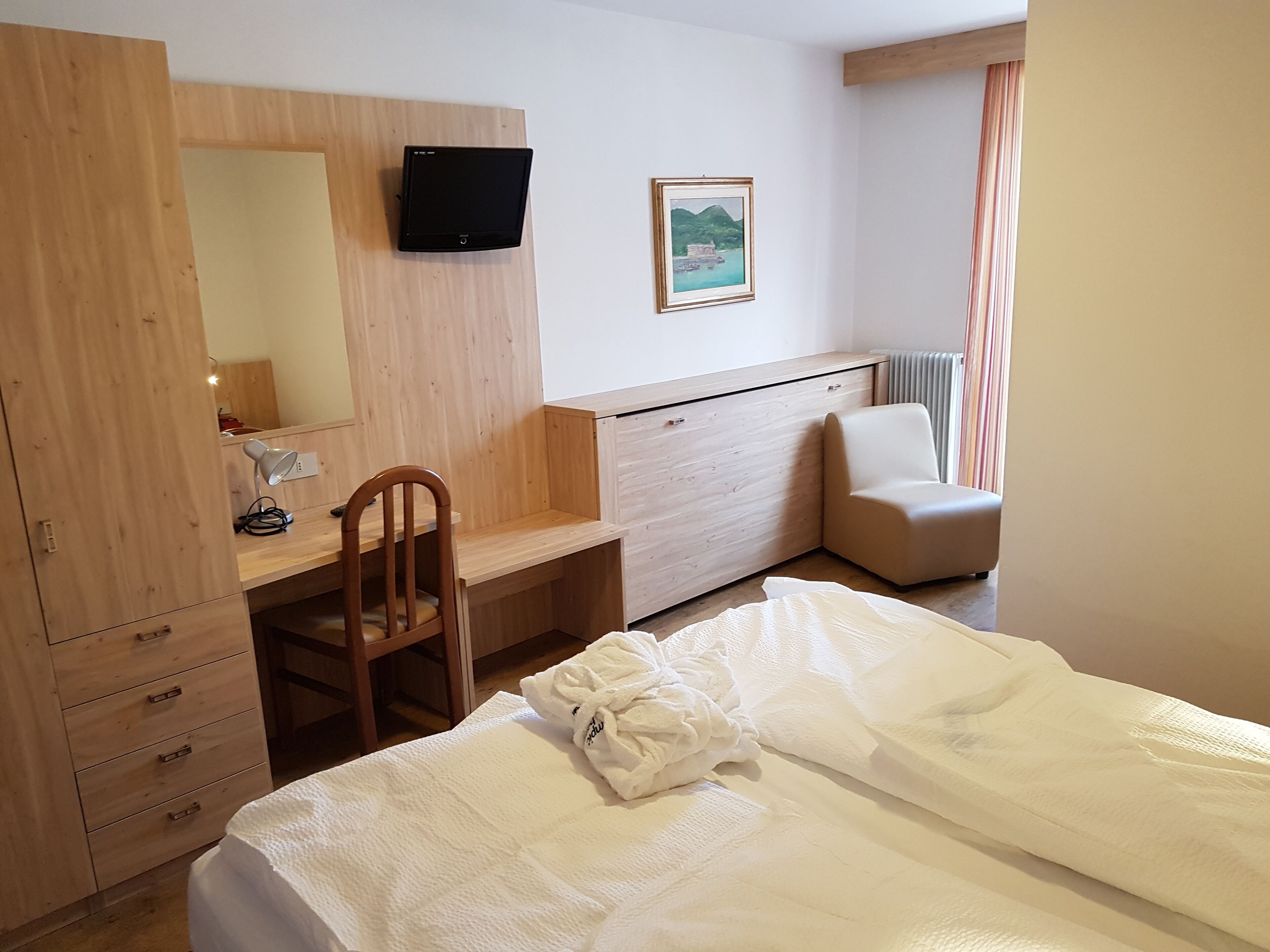 Habitación de hotel con escritorio de madera, una silla, cama con ropa de cama blanca, televisor montado en la pared y ventana con cortinas.
