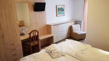 Habitación de hotel con escritorio de madera, una silla, cama con ropa de cama blanca, televisor montado en la pared y ventana con cortinas.