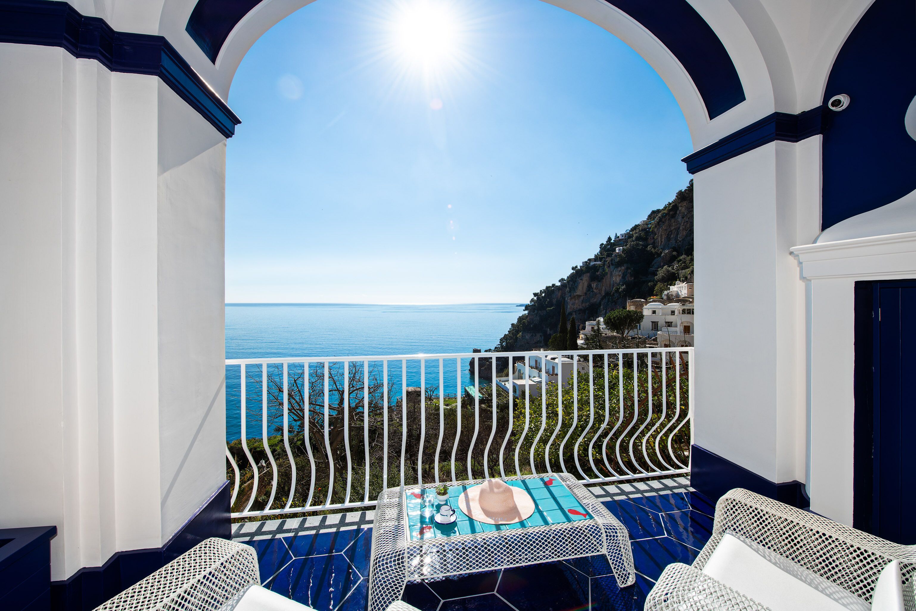 Un balcón con vista al mar, muebles de mimbre blanco y una mesa con un sombrero y una botella.