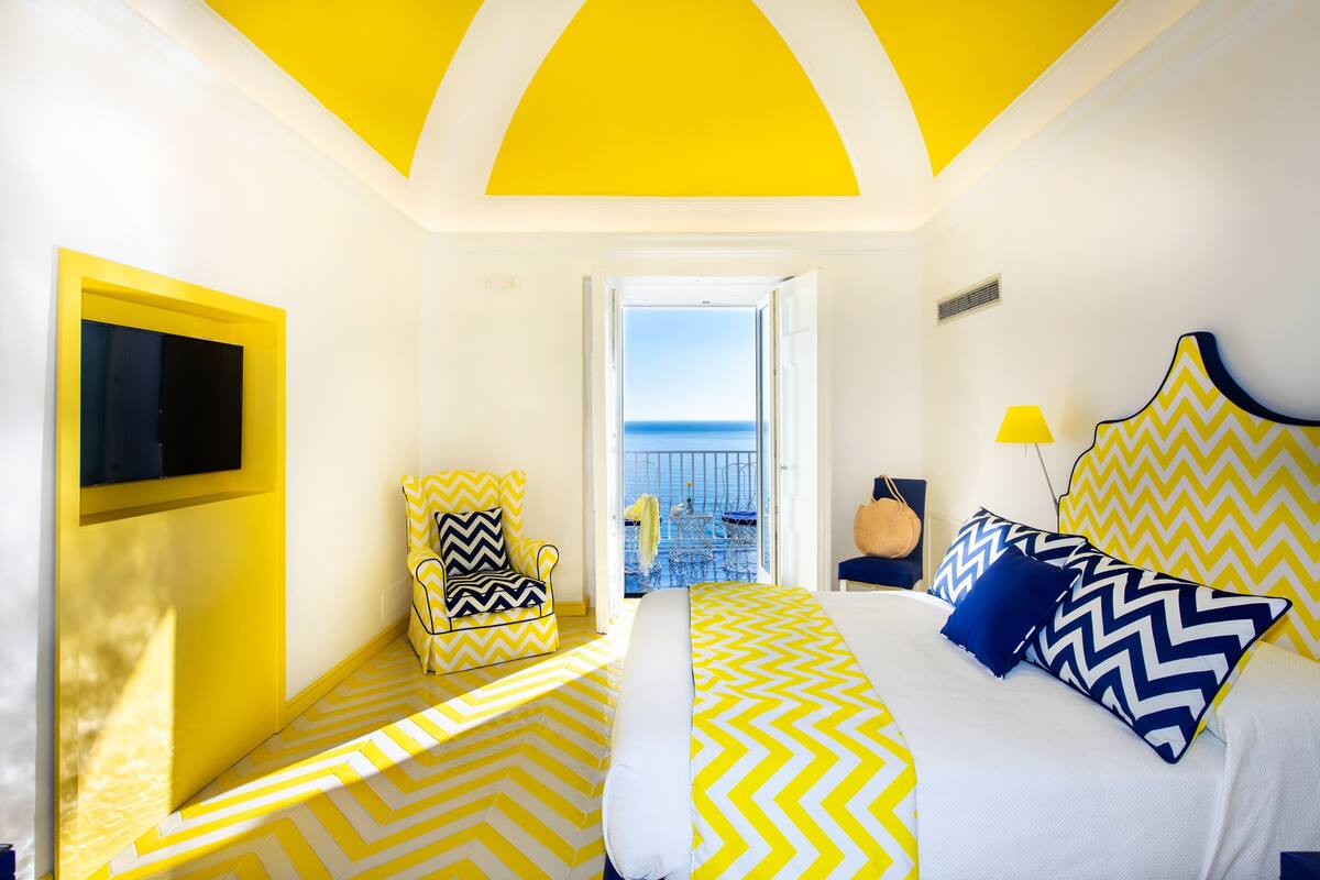 A bright room with yellow and white walls, a bed with patterned bedding, a chair with a zigzag pillow, and a view of the sea through an open door.