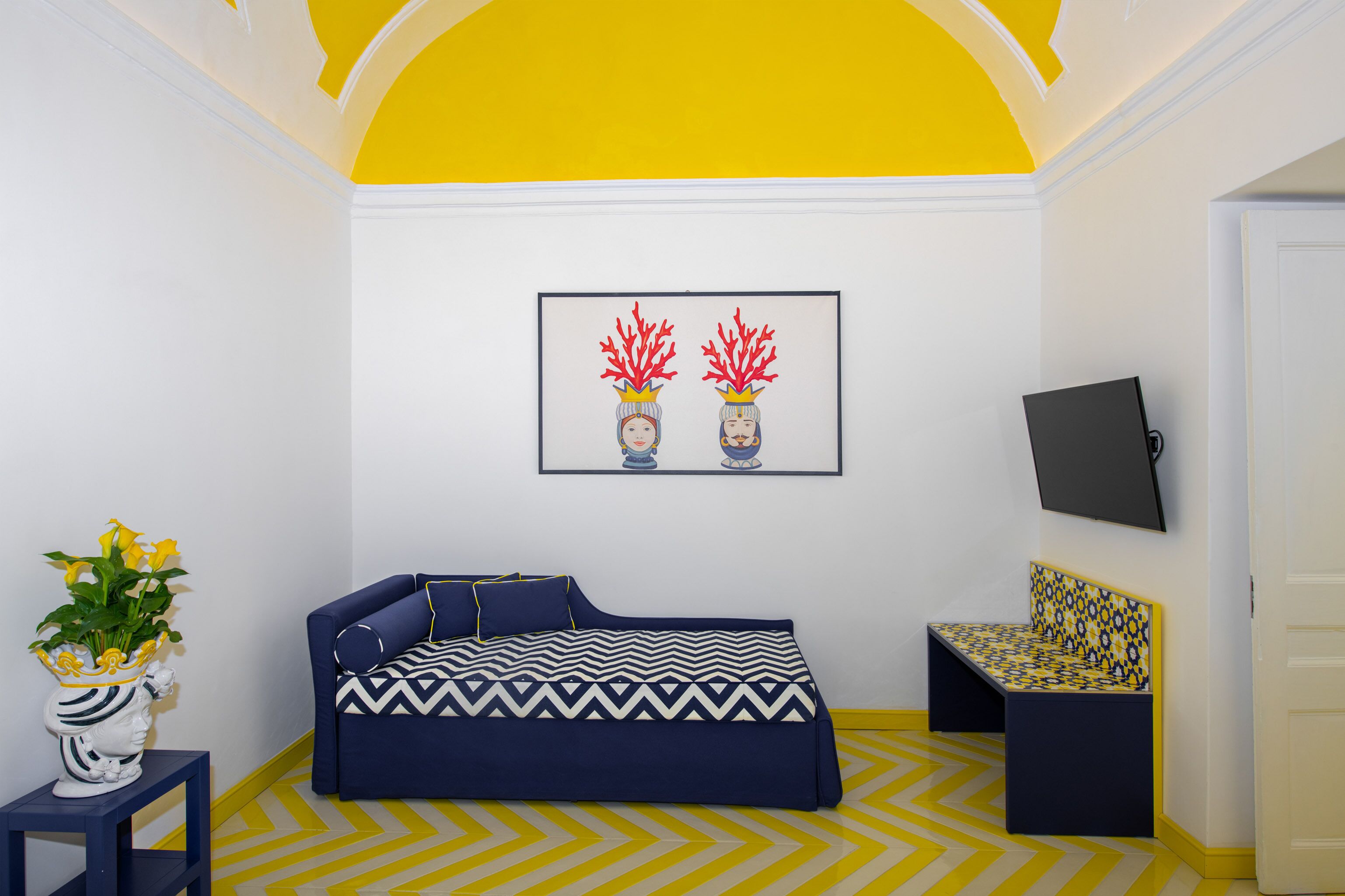 A room with a blue sofa, a small table with a vase of flowers, a patterned bench, and a framed picture on the wall.