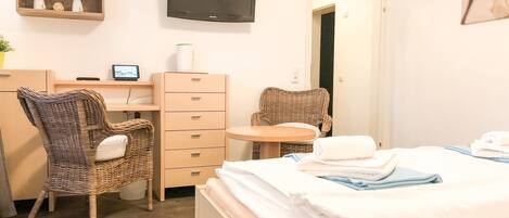 Une chambre d’hôtel comprenant un lit, deux fauteuils en osier, un bureau avec un ordinateur, une petite table ronde et un téléviseur fixé au mur.