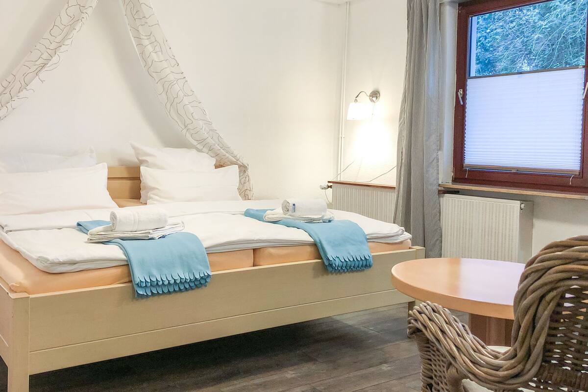 A bed with a canopy, a bedside table, a lamp, a window with curtains, and a wicker chair.