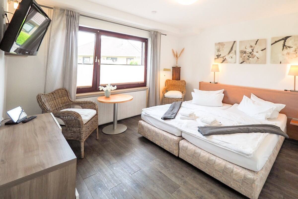 A hotel room with a bed, a chair, a small table, a TV, and artwork on the wall.