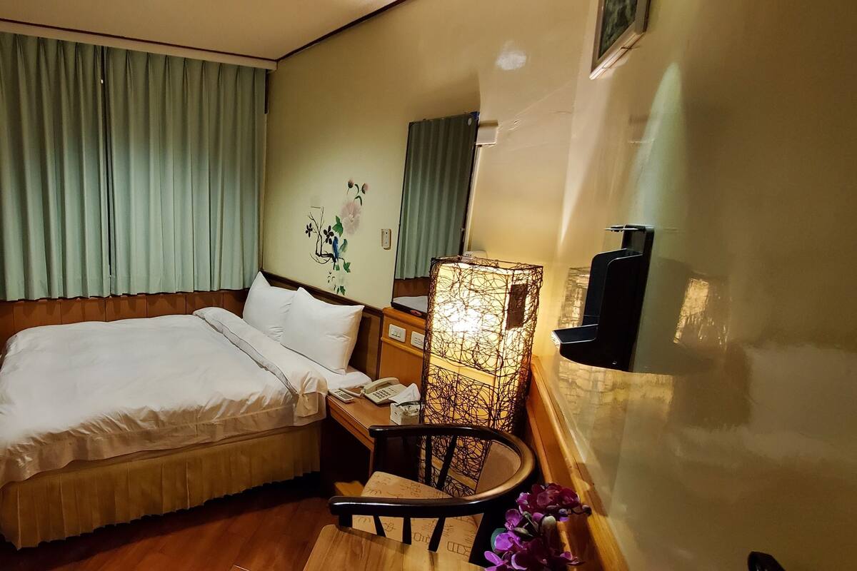 Habitación de hotel con cama, una silla, un escritorio con lámpara, televisión y aire acondicionado instalado en la pared.