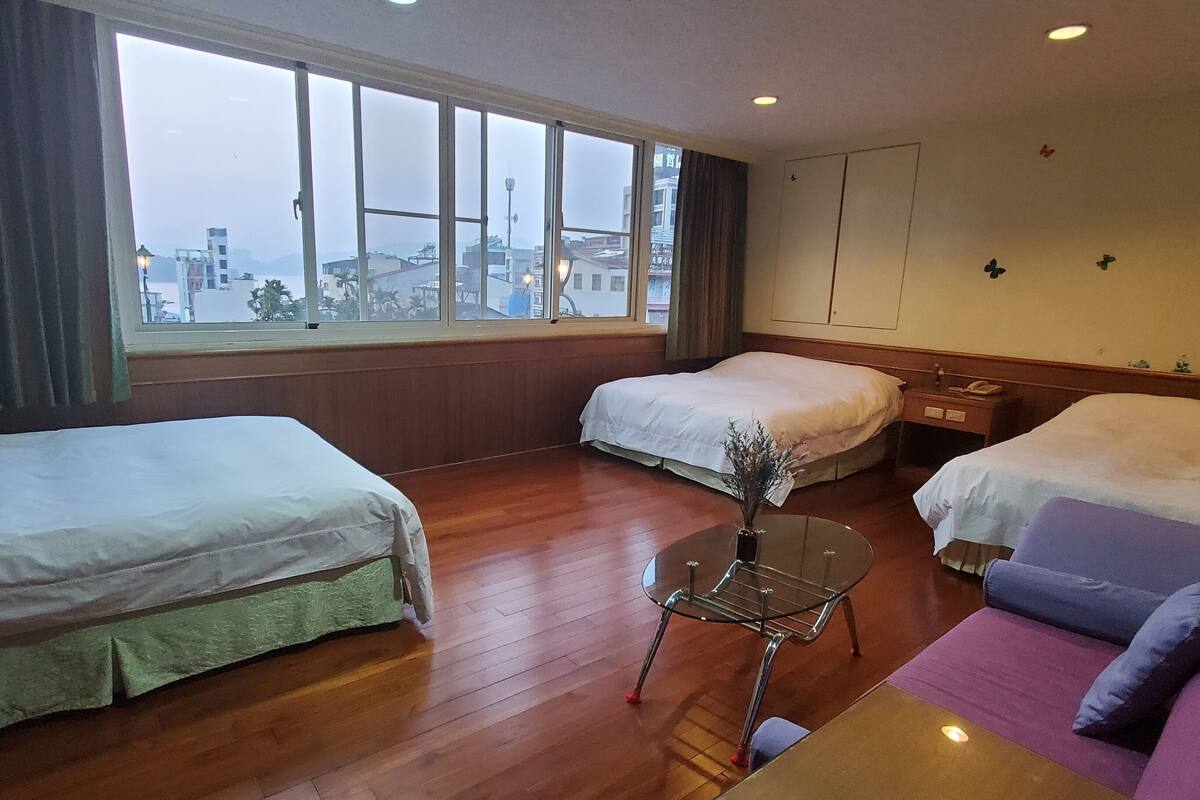 Habitación de hotel con dos camas, un sofá, una mesita y vistas a la ciudad a través de amplias ventanas.