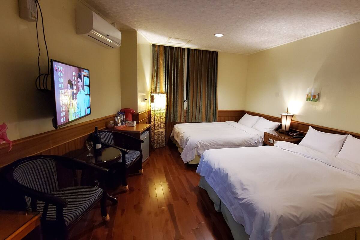 Habitación de hotel con dos camas, televisión, escritorio con silla, florero con flores y lámpara.
