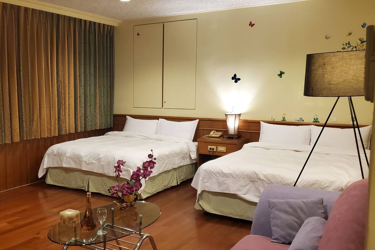 Habitación de hotel con dos camas, una mesita pequeña con un florero, una lámpara y una mesita de noche con un teléfono.