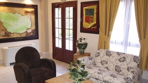 Un salón con sofá, sillón y mesa de centro. Hay un mapa en la pared, una puerta y una ventana con cortinas.