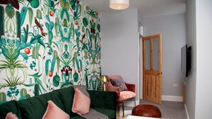 A living room with a green sofa, a patterned wallpaper, a wooden door, and a television mounted on the wall. - The Old Post Office - Bright & Modern 4bdr Townhouse With Private Garden (Oxford)