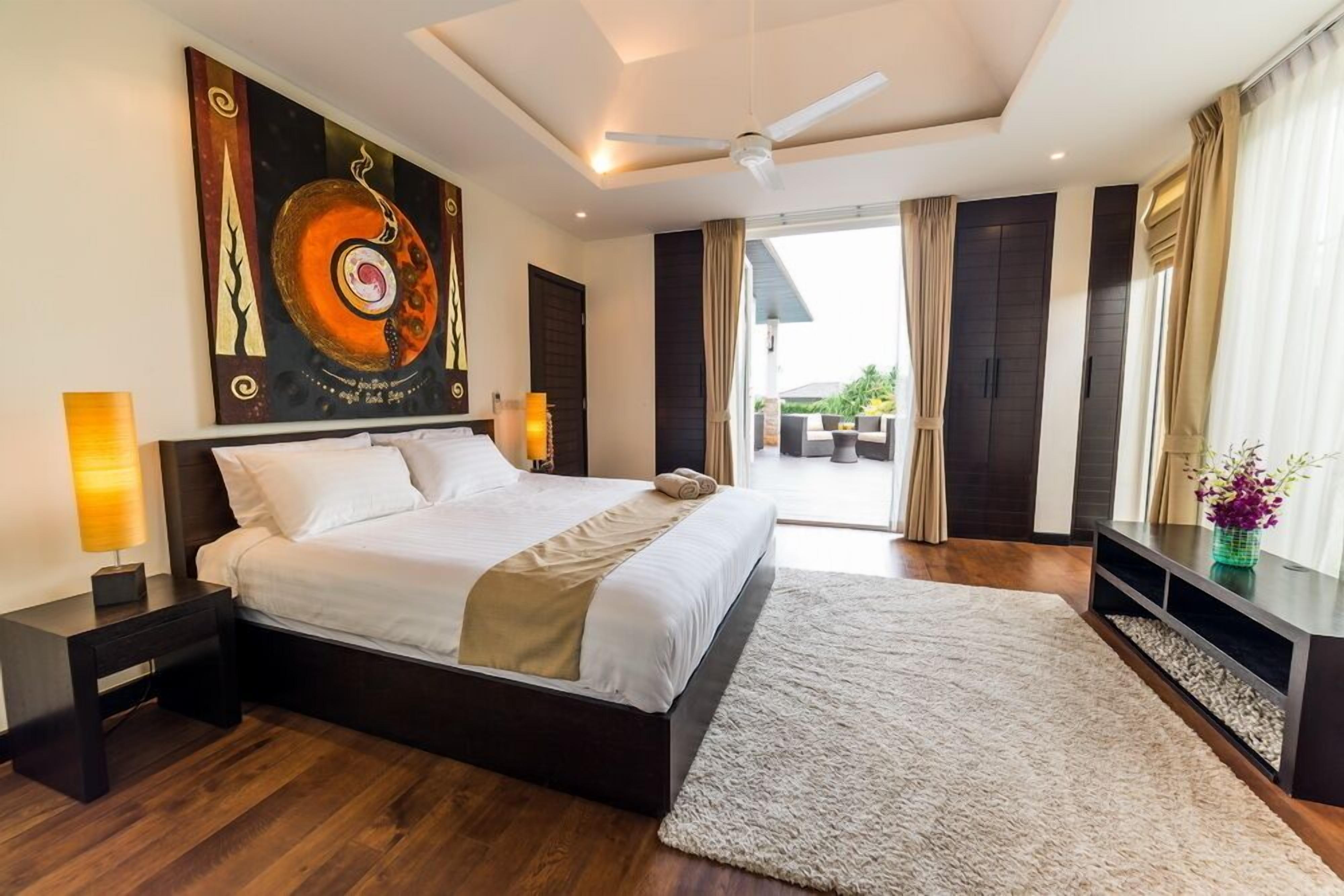 A modern hotel bedroom with a large bed, a ceiling fan, a view of the outdoors, and a painting on the wall.