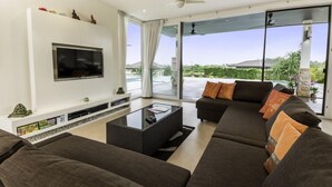 Un salon moderne doté d’un grand téléviseur à écran plat, d’un ensemble de canapés, d’une table basse en verre et offrant une vue sur l’extérieur grâce à de grandes portes-fenêtres coulissantes.