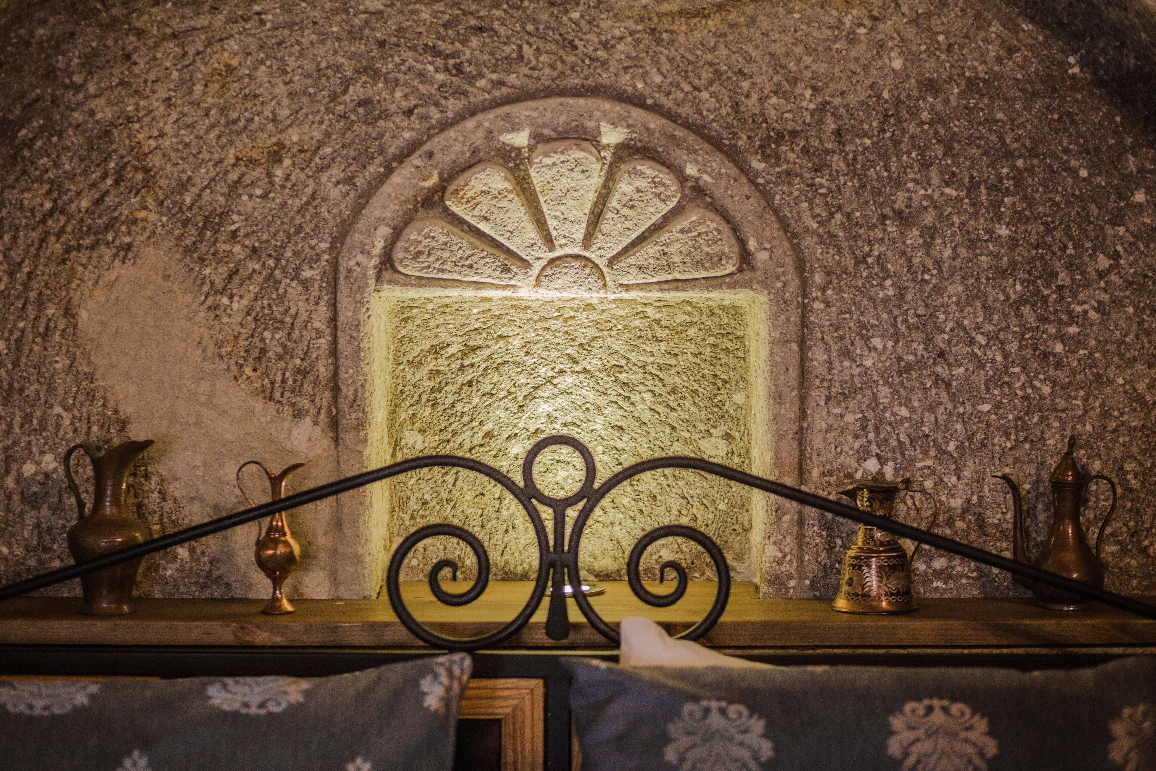 A room with a stone wall, a decorative metal grill, and two metal pitchers on a wooden surface.