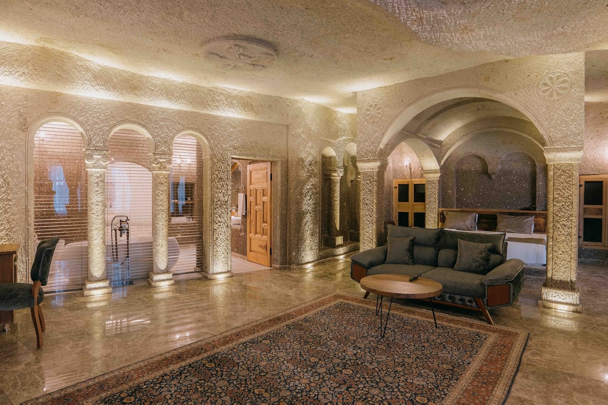 Un vestíbulo espacioso con un sofá grande, una mesa de centro y una alfombra estampada. Se observan múltiples arcos y una puerta de madera que conduce a otra habitación.