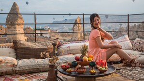 Una persona sentada en una cama con una bandeja de frutas y un tazón de nueces, con vista a un paisaje urbano y globos aerostáticos al fondo.