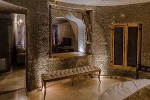 A stone-walled room with a bench, a bed, a mirror, and a window with shutters.