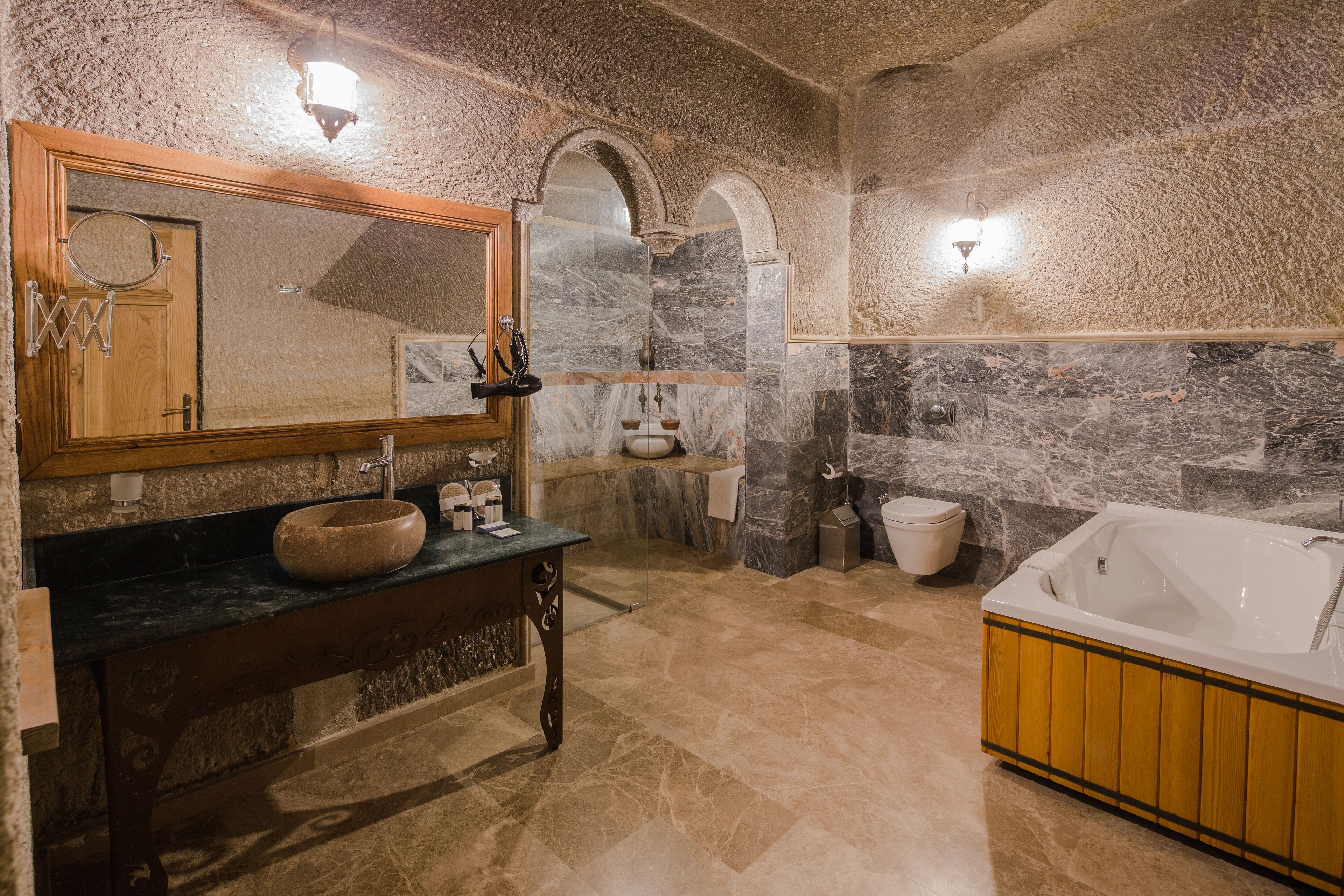 A bathroom with a stone wall, a wooden vanity, a large mirror, a bathtub, and a toilet.