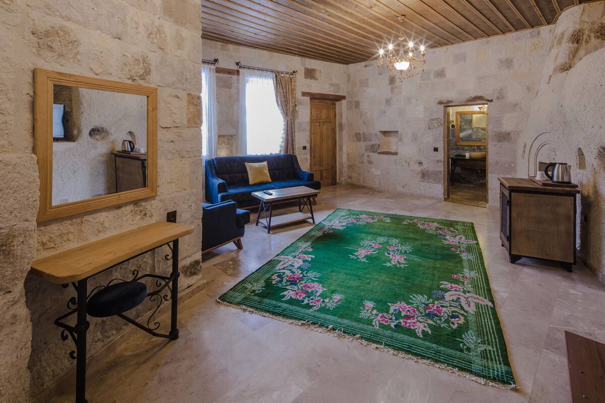 Una habitación rústica con techo de madera, alfombra floral verde, sofá azul, mesa de madera y un espejo en la pared.