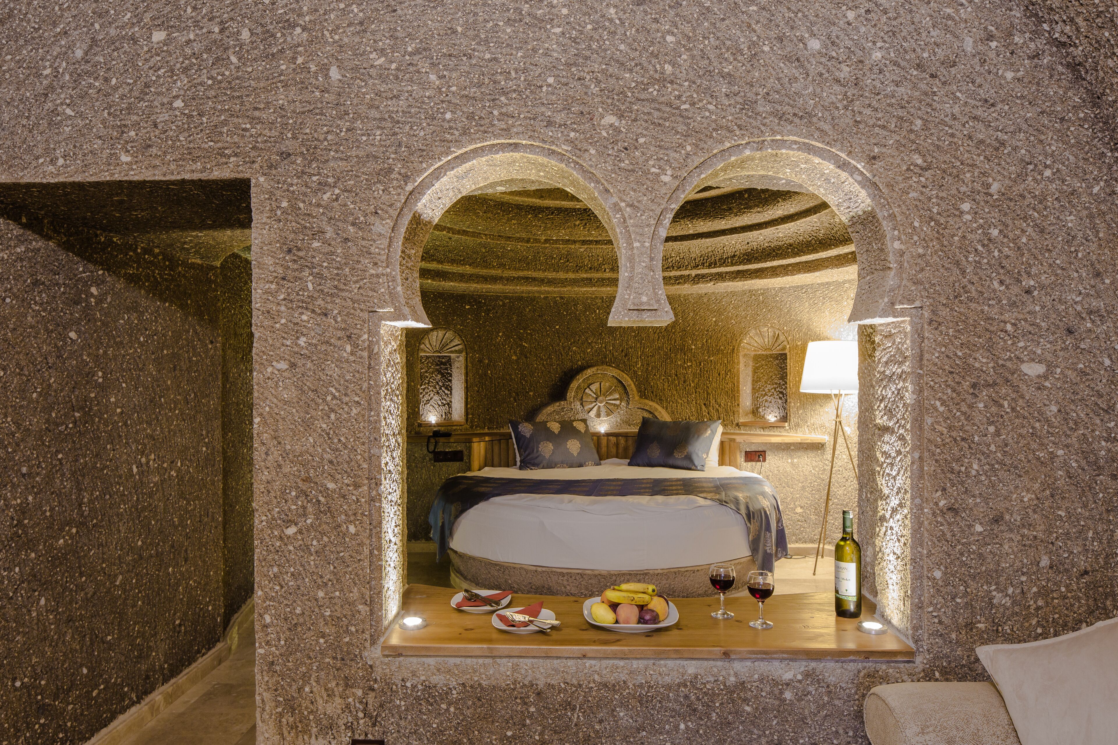 Une chambre aux murs de pierre, avec un lit, une petite table sur laquelle se trouvent de la nourriture et du vin, et une lampe.