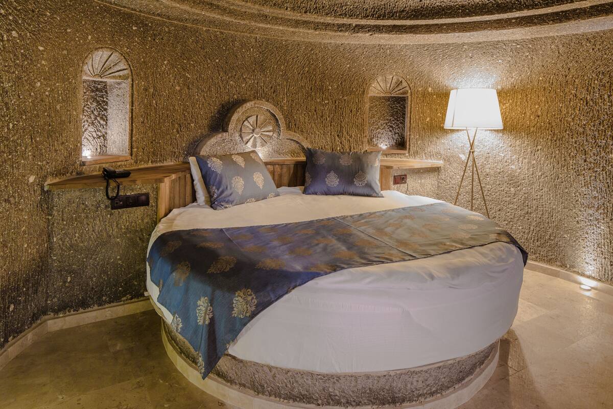 Una habitación circular con una cama centrada, una lámpara sobre un pedestal y una ventana con un ventilador.