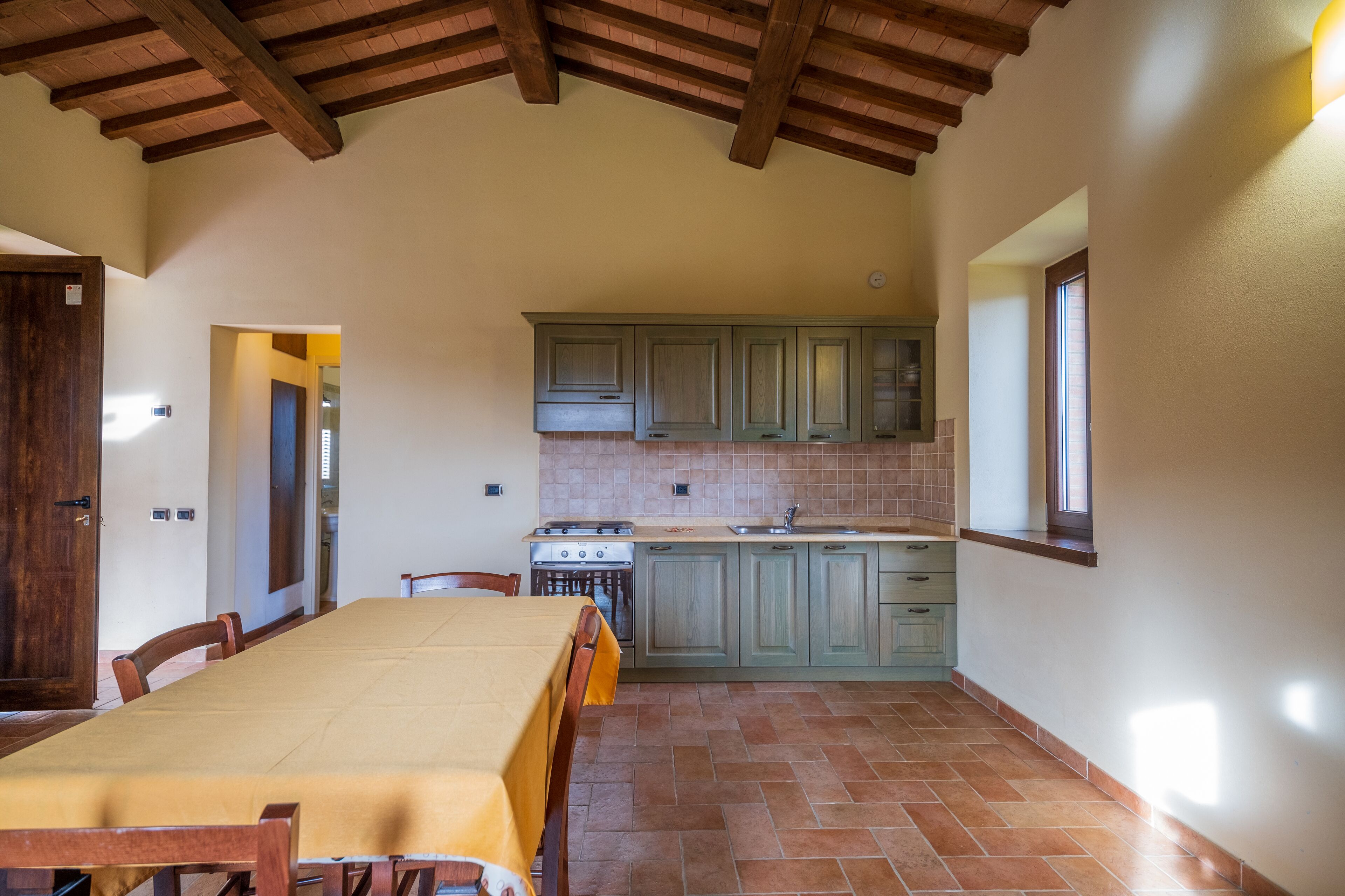 A spacious dining area with a long table and chairs, a kitchen with green cabinets and a stove, and a wooden ceiling.
