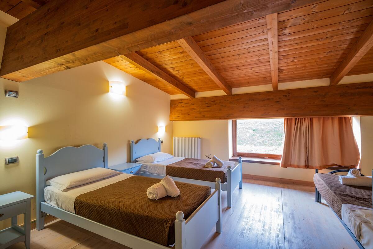 Una habitación con dos camas, vigas de madera en el techo, una ventana con cortinas y una mesita de noche con lámpara.