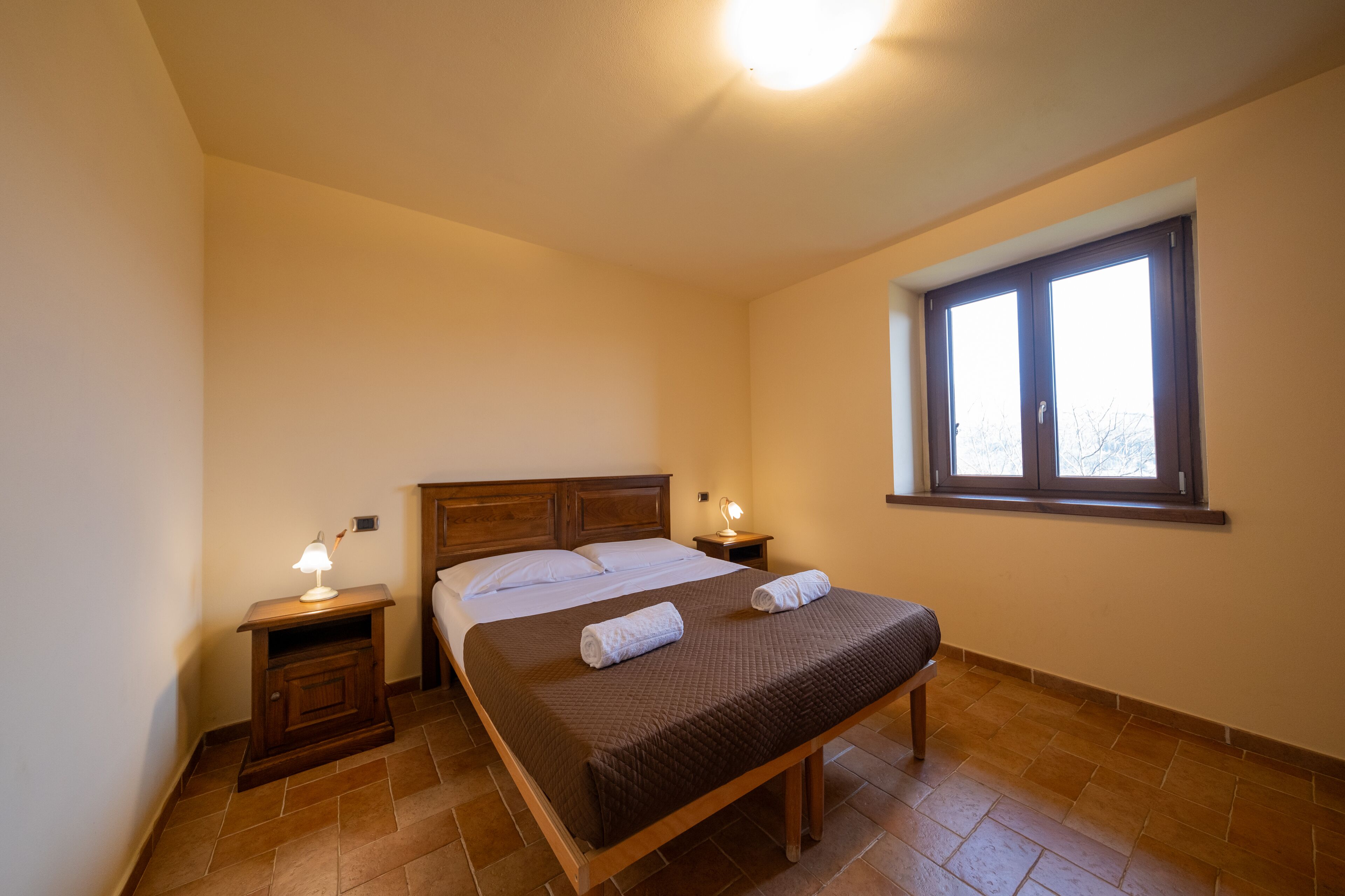A bedroom with a bed, two bedside tables, a window, and a nightstand with a lamp.