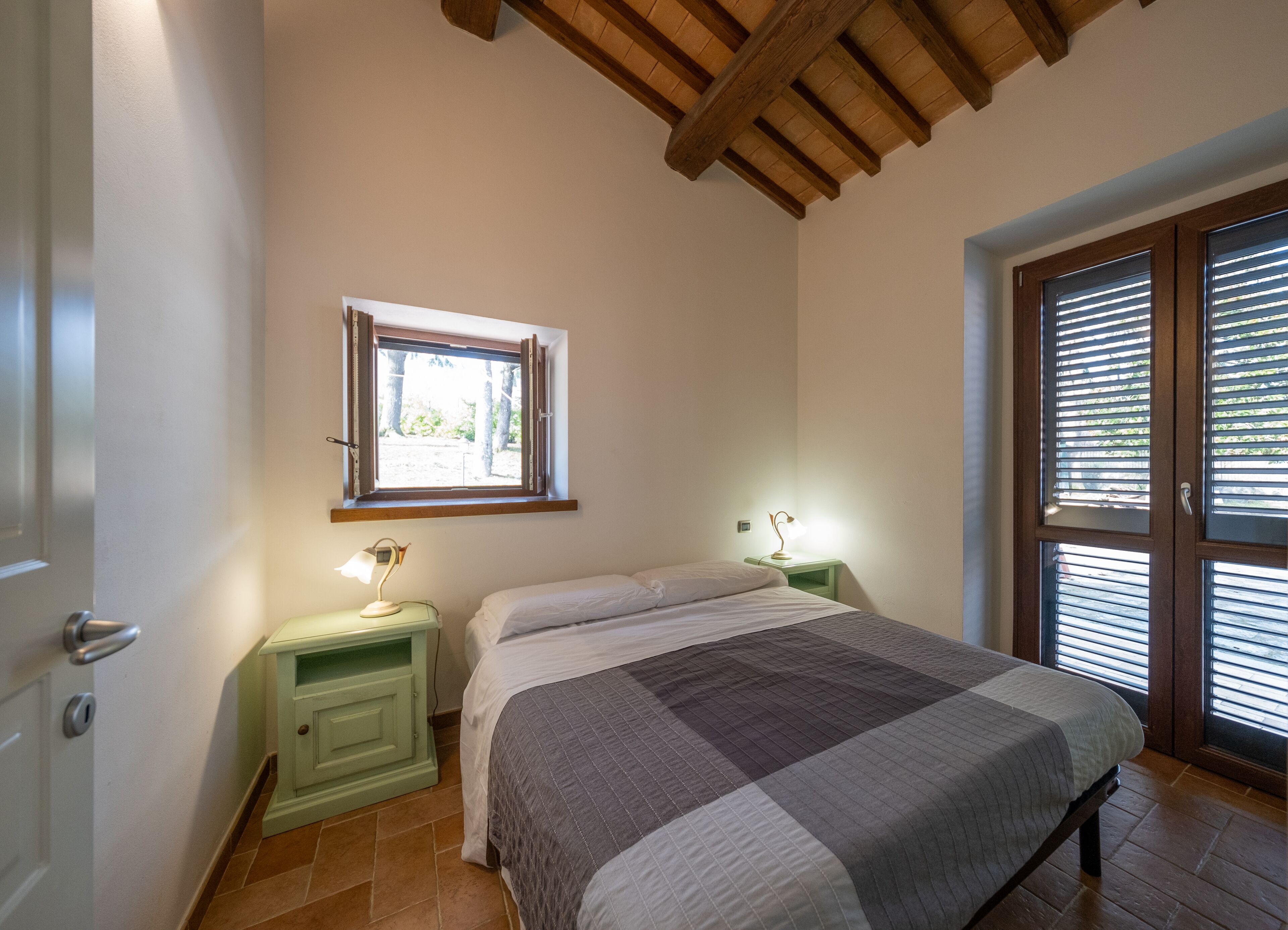 A bedroom with a bed, a nightstand with a lamp, a window with blinds, and a door.