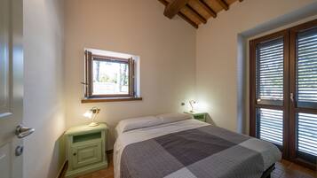 Un dormitorio con cama, una mesita de noche con lámpara, una ventana con persianas y una puerta.