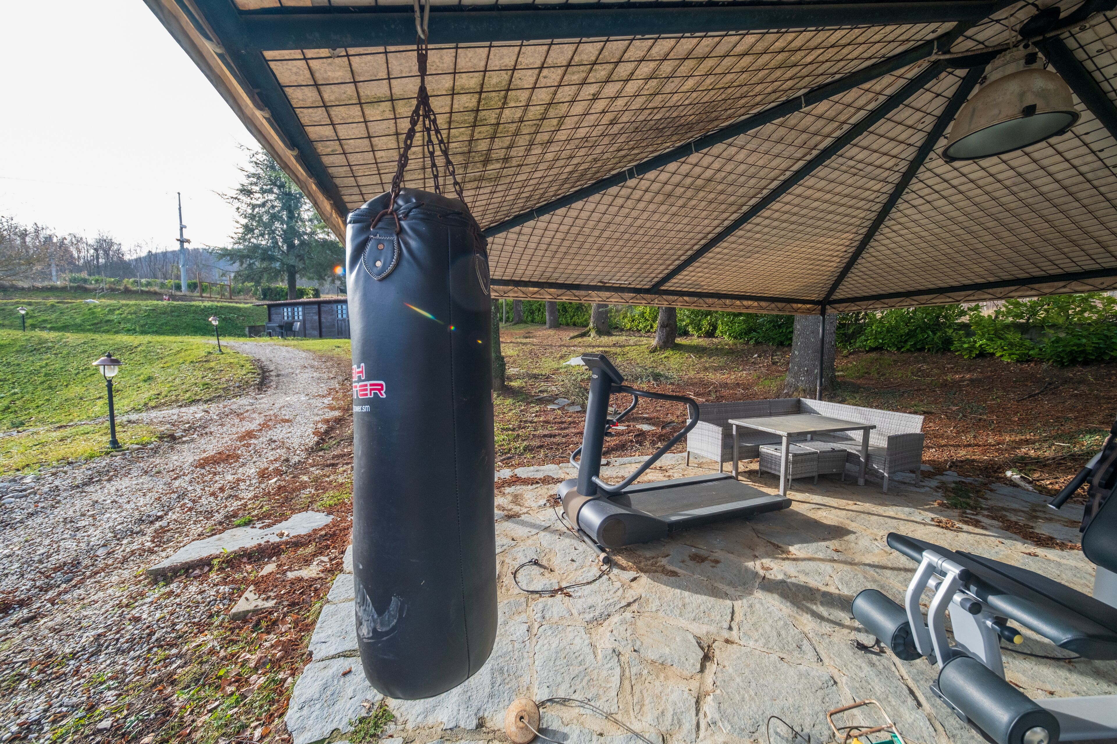 Ein überdachter Trainingsbereich im Freien mit einem Sandsack, Fitnessgeräten und einem Tisch unter einer Markise.