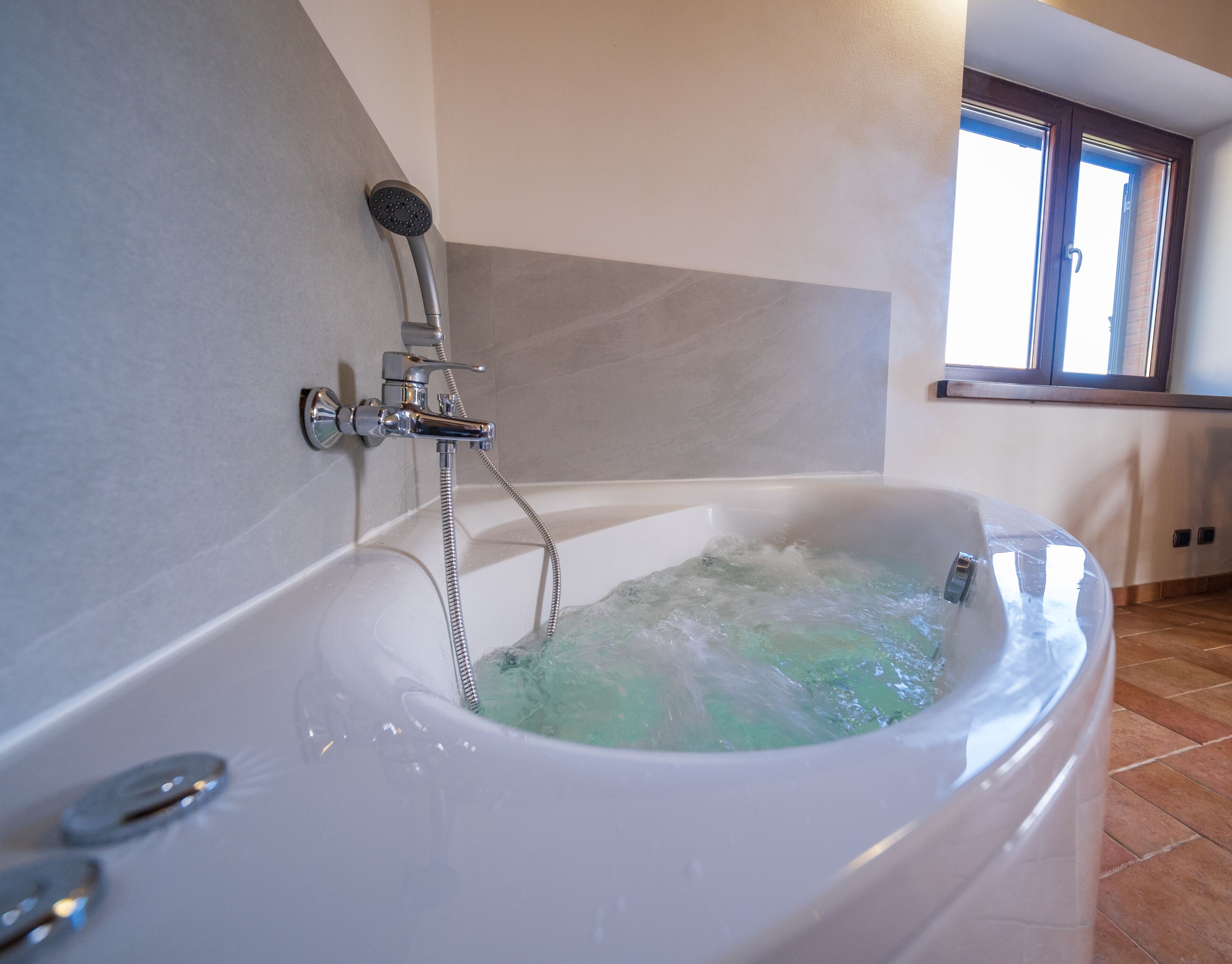 A bathroom with a large bathtub filled with water, a showerhead, and a window providing natural light.