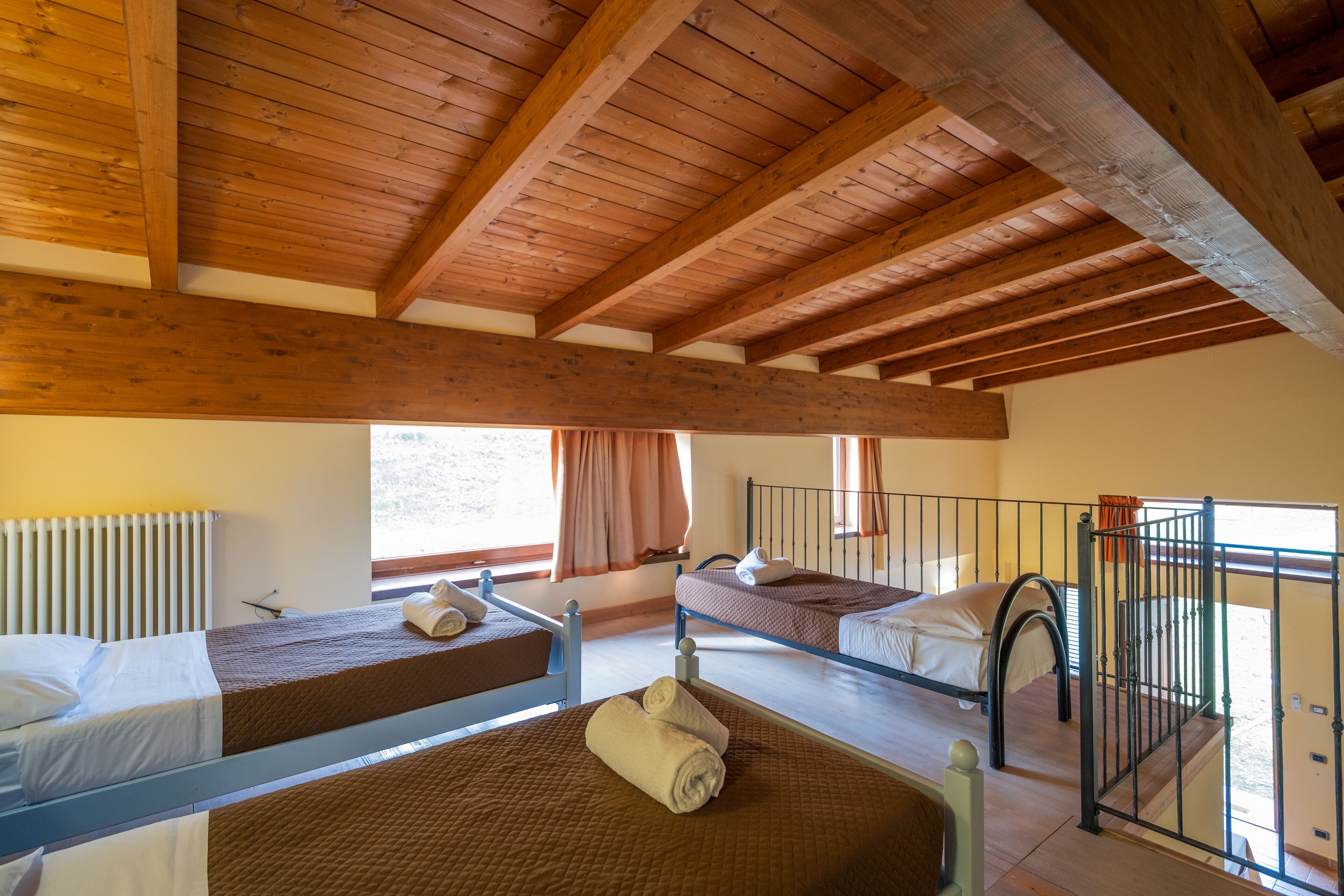 Une pièce avec trois lits superposés, des poutres en bois au plafond, une fenêtre avec des rideaux et un radiateur.